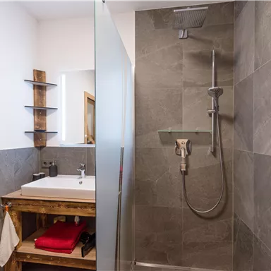 Ein modernes Badezimmer mit einer Dusche und einem schlichten Waschbecken. Die Wände sind mit grauen Fliesen verkleidet und es gibt eine Holzablage.