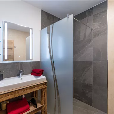 Ein modernes Badezimmer mit einer Dusche und einem Waschbecken aus Holz. Die Wände sind in neutralen Farbtönen gehalten, und es gibt einige rote Handtücher.