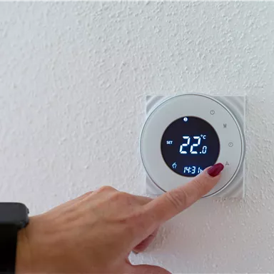 Ein moderner Thermostat an der Wand zeigt eine Temperatur von 22°C an. Eine Hand mit einem Nagellack drückt auf die Steuerelemente.