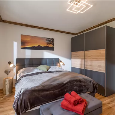 Ein modernes Schlafzimmer mit einem großen Bett und einem eleganten Kleiderschrank. Rote Handtücher liegen auf einer Sitzbank am Fußende des Bettes.