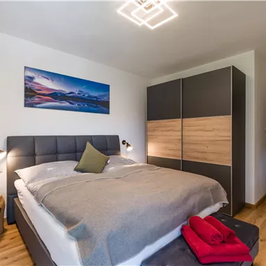 Ein modernes Schlafzimmer mit einem großen Doppelbett und stilvollen Möbeln. An der Wand hängt ein Bild von einer Landschaft und der Boden ist mit Holz ausgelegt.