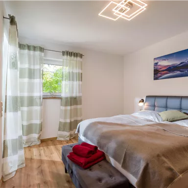 Ein gemütliches Schlafzimmer mit einem Doppelbett, einem großen Fenster und hellen Vorhängen. Ein modernes Bild hängt an der Wand und auf dem Bett liegen eine Decke und rotes Handtuch.