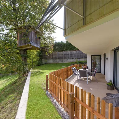 Eine gemütliche Terrasse mit Gartenmöbeln, umgeben von einem gepflegten Garten. Im Hintergrund steht ein Baumhaus, das an einen großen Baum angelehnt ist.