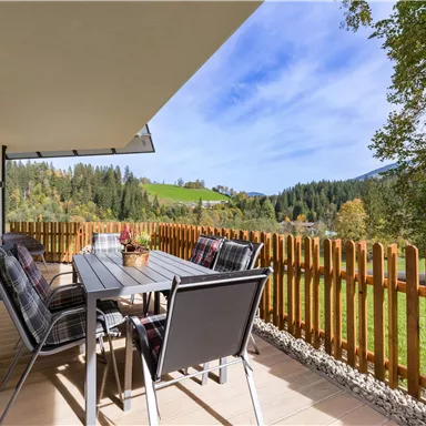Eine gemütliche Terrasse mit einem Esstisch und Stühlen. Im Hintergrund ist eine malerische Landschaft mit grünen Hügeln und Bäumen zu sehen.
