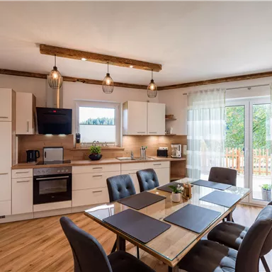 Eine moderne Küche mit einem Esstisch und Stühlen. Große Fenster lassen viel Licht herein und bieten einen Blick nach draußen.