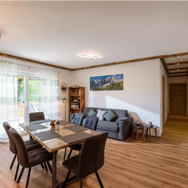 Ein modernes Wohnzimmer mit einem Esstisch und gemütlichem Sofa. Große Fenster lassen viel Licht herein und der Boden ist aus Holz.