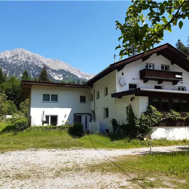 A beautiful building amidst green meadows and trees, with majestic mountains in the background. The sky is clear and blue.