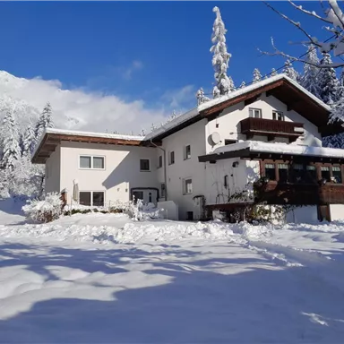 A charming house in a winter landscape, surrounded by snow and tall trees. The clear blue sky highlights the tranquil atmosphere.