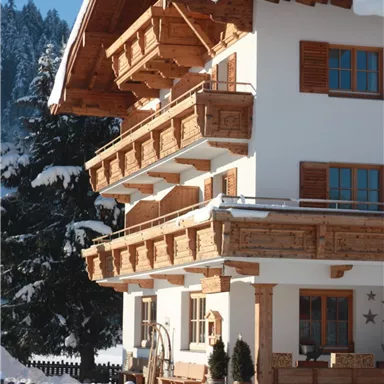 Ein malerisches Haus im Alpenstil, umgeben von Schnee. Die Sonne scheint auf das Holzgebäude und die angrenzende Landschaft.