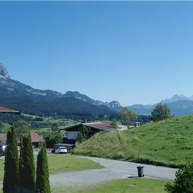 Eine ruhige Landschaft mit Bergen im Hintergrund und klar blauem Himmel. Im Vordergrund sind Häuser und grüne Wiesen zu sehen.