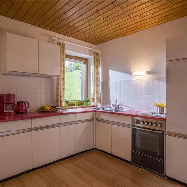 Eine moderne Küche mit weißen Schränken und Holzdecken. Licht fällt durch das Fenster und es gibt verschiedene Geräte sowie eine Arbeitsfläche.