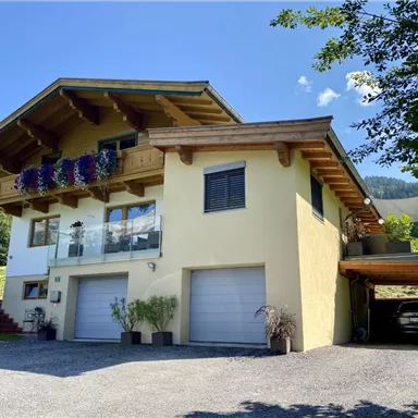 A modern house with a large balcony and flower boxes. The parking area is covered and there is a green landscape in the background.