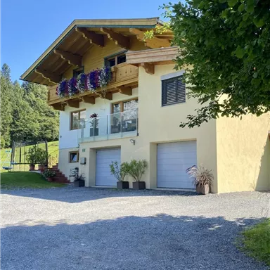 A modern house with a yellow paint and a balcony full of flowers. It is located in a green environment, surrounded by trees.