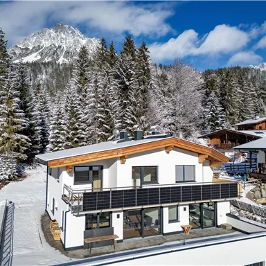 A modern house in a snowy landscape, surrounded by tall fir trees. In the background, majestic mountains and a bright blue sky can be seen.