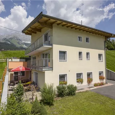 A modern house in a green environment with a sunny garden seating area. In the background, mountains and a clear sky can be seen.