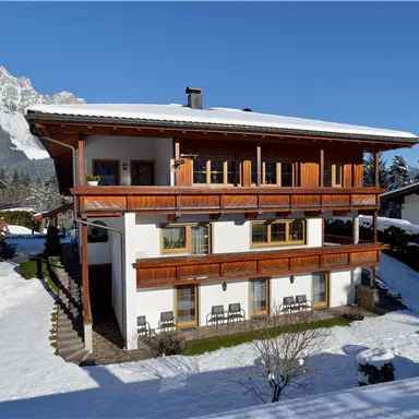 Ein charmantes Haus im Schnee mit einem großen Balkon und Holzverzierungen. Im Hintergrund sind schneebedeckte Berge und Bäume zu sehen.