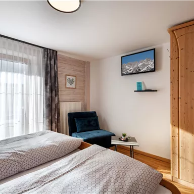 Ein gemütliches Schlafzimmer mit einem Doppelbett, einem Sessel und einem kleinen Tisch. An der Wand hängt ein Fernseher und es gibt große Fenster mit Vorhängen.