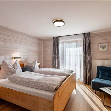 Ein gemütliches Schlafzimmer mit zwei Betten und holzernen Möbeln. Ein blauer Sessel und ein Fenster mit Vorhängen bieten einen einladenden Ausblick.