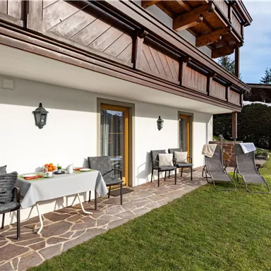 Eine gemütliche Terrasse mit Gartenmöbeln und einem Tisch, der für ein Frühstück gedeckt ist. Im Hintergrund sind grüne Wiesen und Bäume zu sehen.