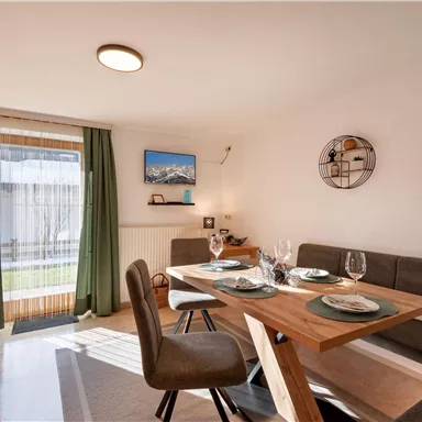 Ein helles Esszimmer mit einem holzfarbenen Tisch und bequemen Stühlen. Die Fenster lassen viel Licht herein und bieten einen Blick auf den Außenbereich.