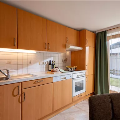 Eine moderne Küche mit Holzschränken und einer hellen Arbeitsfläche. Das Fenster lässt viel Tageslicht herein und bietet Aussicht auf den Außenbereich.