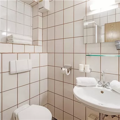 Ein kleines, modernes Badezimmer mit weißen Fliesen. Es verfügt über ein Waschbecken, eine Dusche und frische Handtücher.