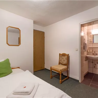 Ein einfaches Schlafzimmer mit einem Einzelbett, einem Stuhl und einem kleinen Waschraum. Die Wände sind weiß und der Raum ist hell und gut beleuchtet.