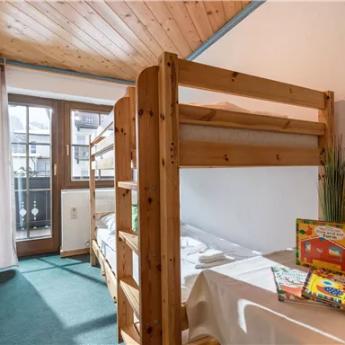 Ein gemütliches Zimmer mit einem Etagenbett und einem Tisch. Es gibt Bücher und einen kleinen Balkon mit Blick auf die Umgebung.