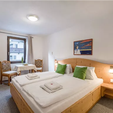 Ein gemütliches Schlafzimmer mit zwei Betten, grünem Dekor und einem Tisch mit Stühlen. Das Zimmer hat große Fenster, die für viel Tageslicht sorgen.