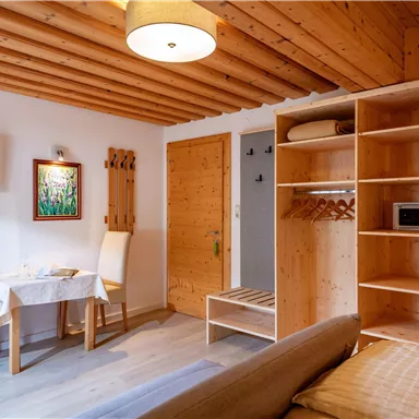A cozy living space with wooden ceiling and walls. There is a table with chairs, a shelf, and access to another room.