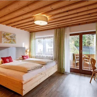 A cozy bedroom with two beds and light wood decor. Large windows offer a view of the balcony and the adjacent nature.