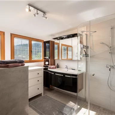 A modern bathroom with large windows and plenty of natural light. The elegant furnishings include a shower, a sink, and a cabinet.