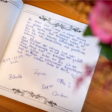An open book with handwritten notes and signatures. In the background, pink flowers can be seen.