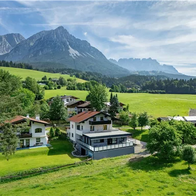 A picturesque landscape with green meadows and mountains in the background. Several houses are set up nearby, surrounded by trees.