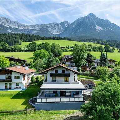 A picturesque landscape with several houses, surrounded by green meadows and trees. In the background, majestic mountains rise under a clear sky.