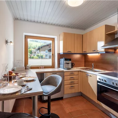 A modern kitchen with wooden cabinets and a large window. The table is set and a bar stool is ready.