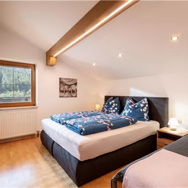 A cozy bedroom with a large bed and a single bed. There is a window with a view outside and warm lighting.