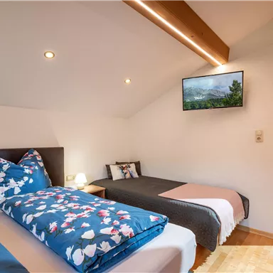 A cozy bedroom with two beds, a wall-mounted television, and warm lighting. The bedding is colorful with floral patterns, and the room feels inviting.