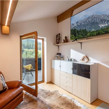 A cozy living room with a leather armchair and a television displaying a mountain landscape. The large window opening provides plenty of daylight and access to the balcony.