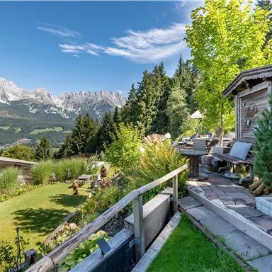A picturesque garden with a wooden terrace overlooking impressive mountains. The landscape is green and sunny, ideal for relaxing.