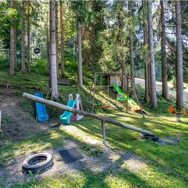 A playground in the forest with slides, swings, and a playhouse. The surroundings are green and filled with light.