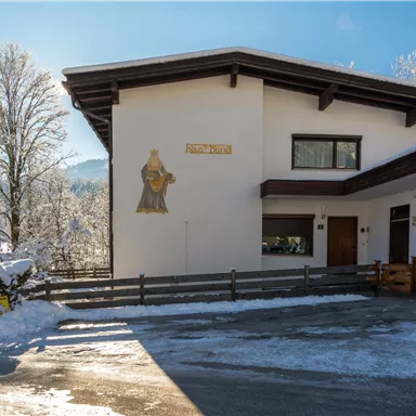 Ein modernes Haus im Winter mit Schnee auf dem Boden. Im Hintergrund sind schneebedeckte Bäume und ein klarer blauer Himmel zu sehen.