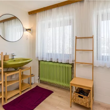 Ein modernes Badezimmer mit einem grünen Waschbecken und einem großen Spiegel. Die Fenster sind mit Vorhängen geschmückt und es gibt einen Regalschrank mit Handtüchern.