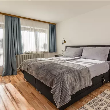 Ein modernes Schlafzimmer mit einem großen Bett und einer gemütlichen Bettwäsche. Große Fenster sorgen für viel Tageslicht und eine helle Atmosphäre.