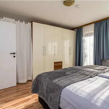 Ein helles Schlafzimmer mit einem großen Bett und Holzfußboden. Vorhänge und ein Kleiderschrank sind ebenfalls sichtbar.