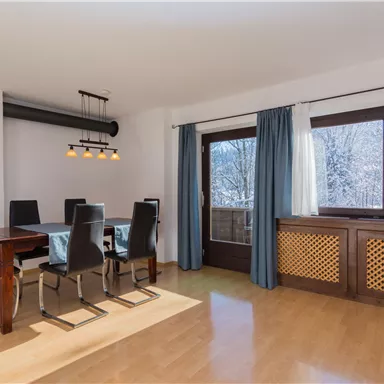 Ein moderner Essbereich mit einem Tisch und sechs Stühlen. Große Fenster lassen viel Tageslicht herein.