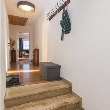 Ein heller Flur mit Holztreppe und moderner Beleuchtung. An der Wand hängen einige Garderobenhaken und ein graues Sitzmöbel steht neben der Treppe.