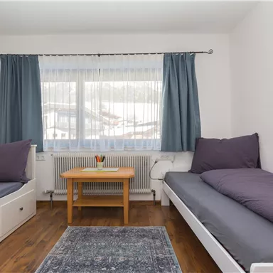 Ein helles Zimmer mit zwei Einzelbetten und einem kleinen Tisch. Große Fenster mit Vorhängen lassen viel Licht herein.