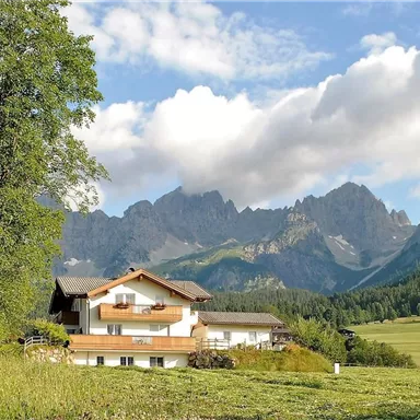 Ein malerisches Haus inmitten der Natur, umgeben von grünen Wiesen und majestätischen Bergen. Der Himmel ist teilweise bewölkt, was eine ruhige Atmosphäre schafft.