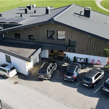 An elegant wooden house with a large roof and colorful flowers in front of the facade. In the foreground, there are parking spaces and a green landscape in the background.